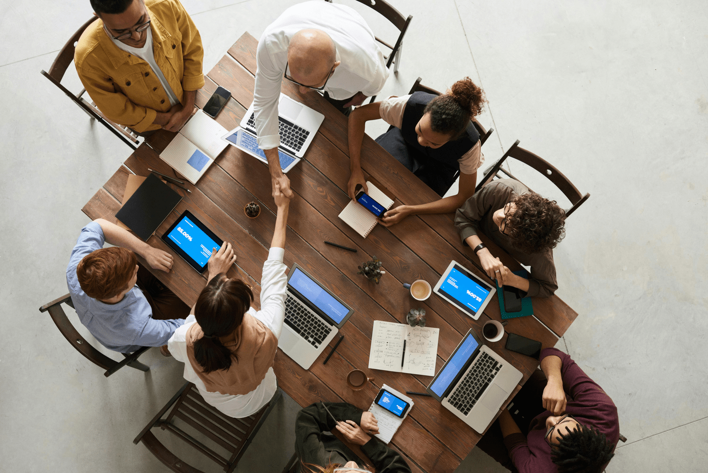 An overhead view of a group of professionals collaborating around a wooden table with laptops, tablets, notebooks, and coffee. Two people are shaking hands while others engage with their devices and notes.